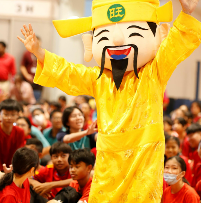 A person in a yellow Chinese deity mascot costume waves at a crowd of smiling children in red clothing.